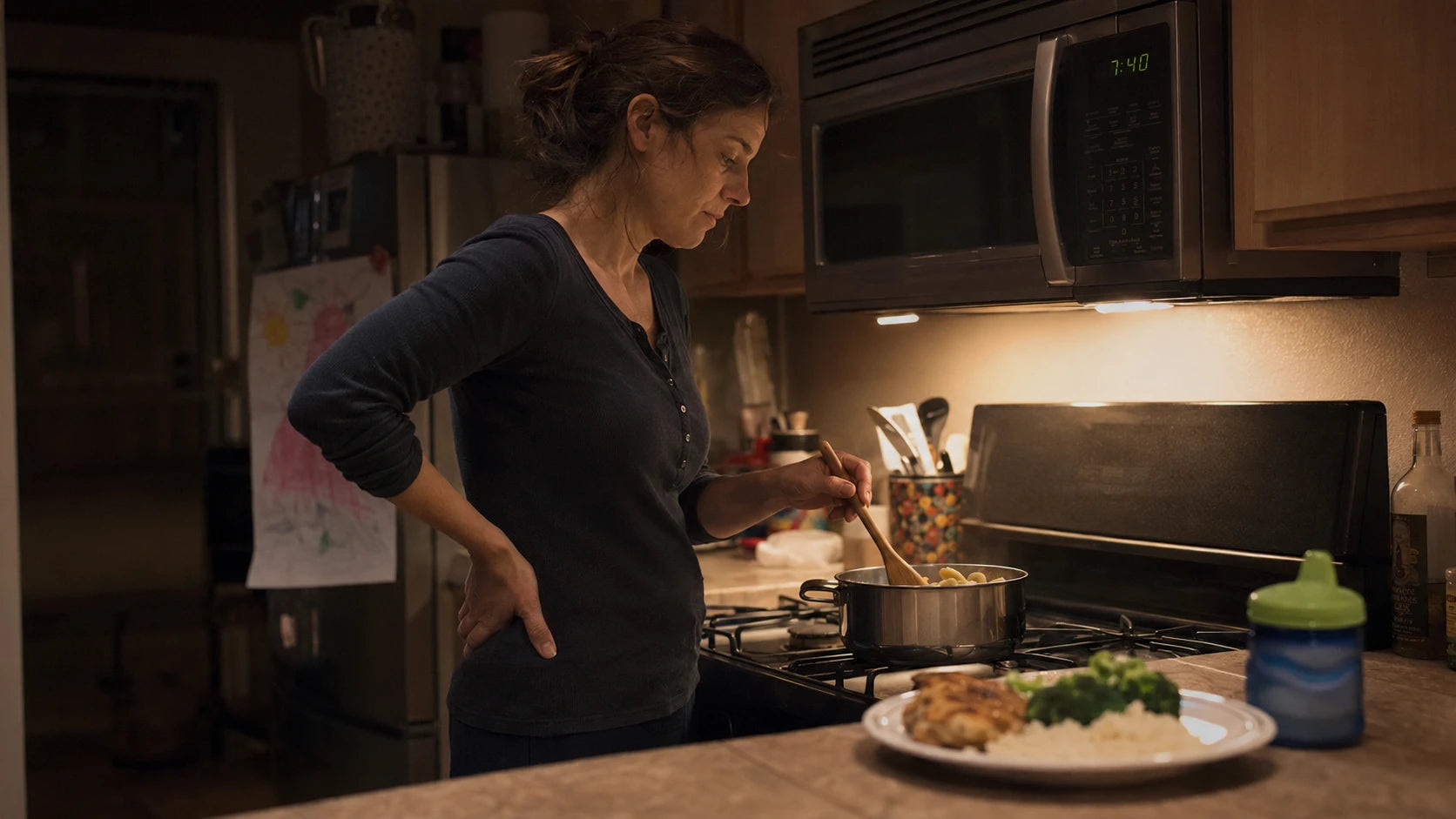 A mother alone at the stove late in the evening, cooking a second small meal while the first rejected plate sits cold on the counter
