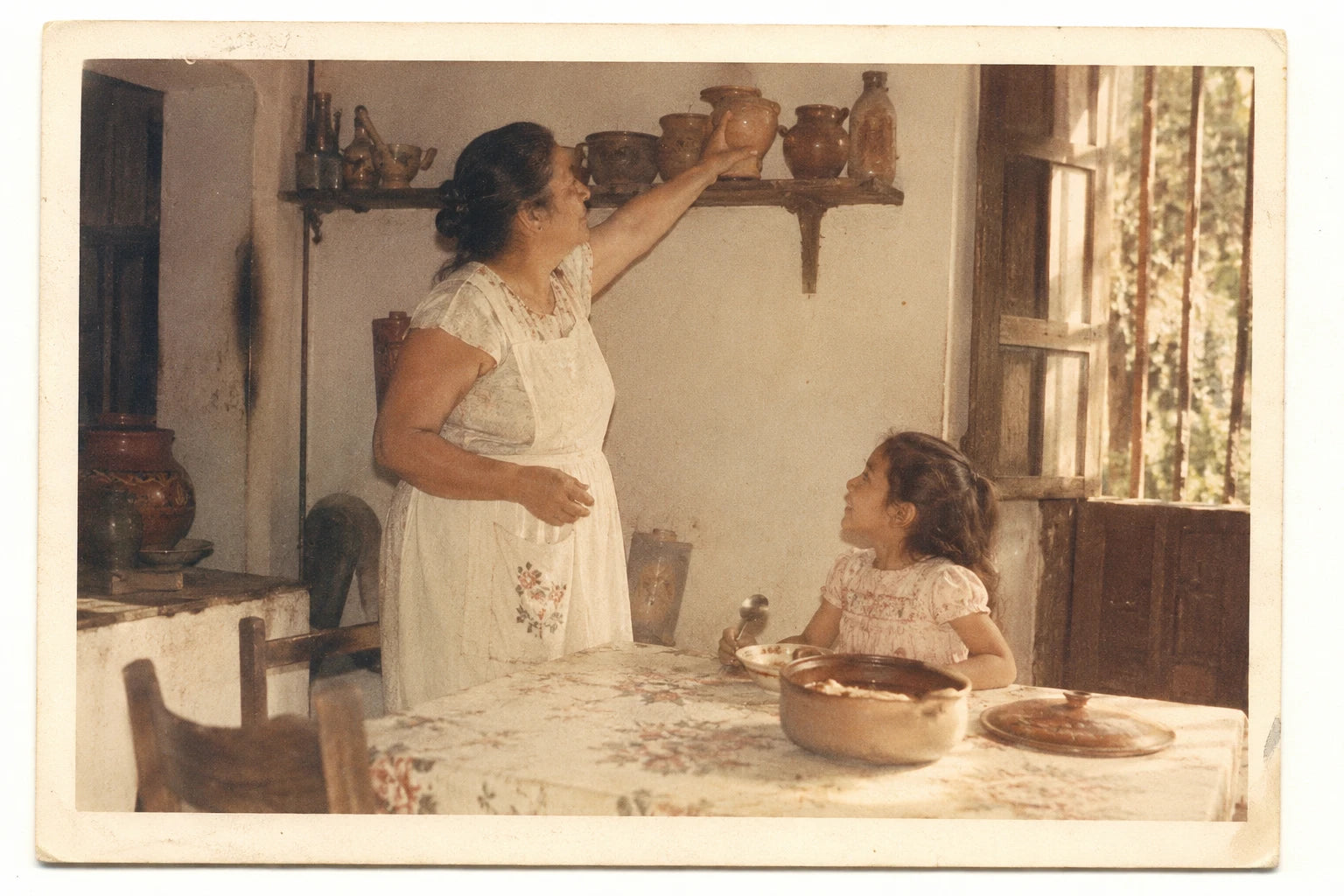 A faded 1960s family photo: a grandmother lifting a small clay pot down in a sunlit Yucatan kitchen while a little girl waits with a spoon
