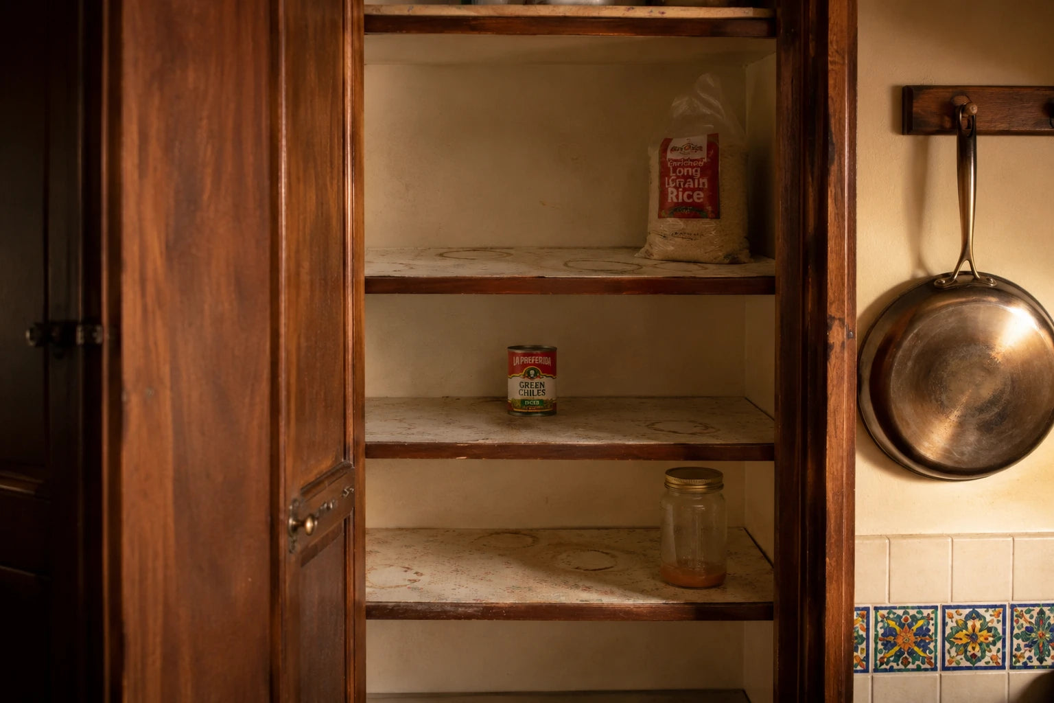 An open kitchen pantry cupboard, nearly empty, with a good pan hanging beside it